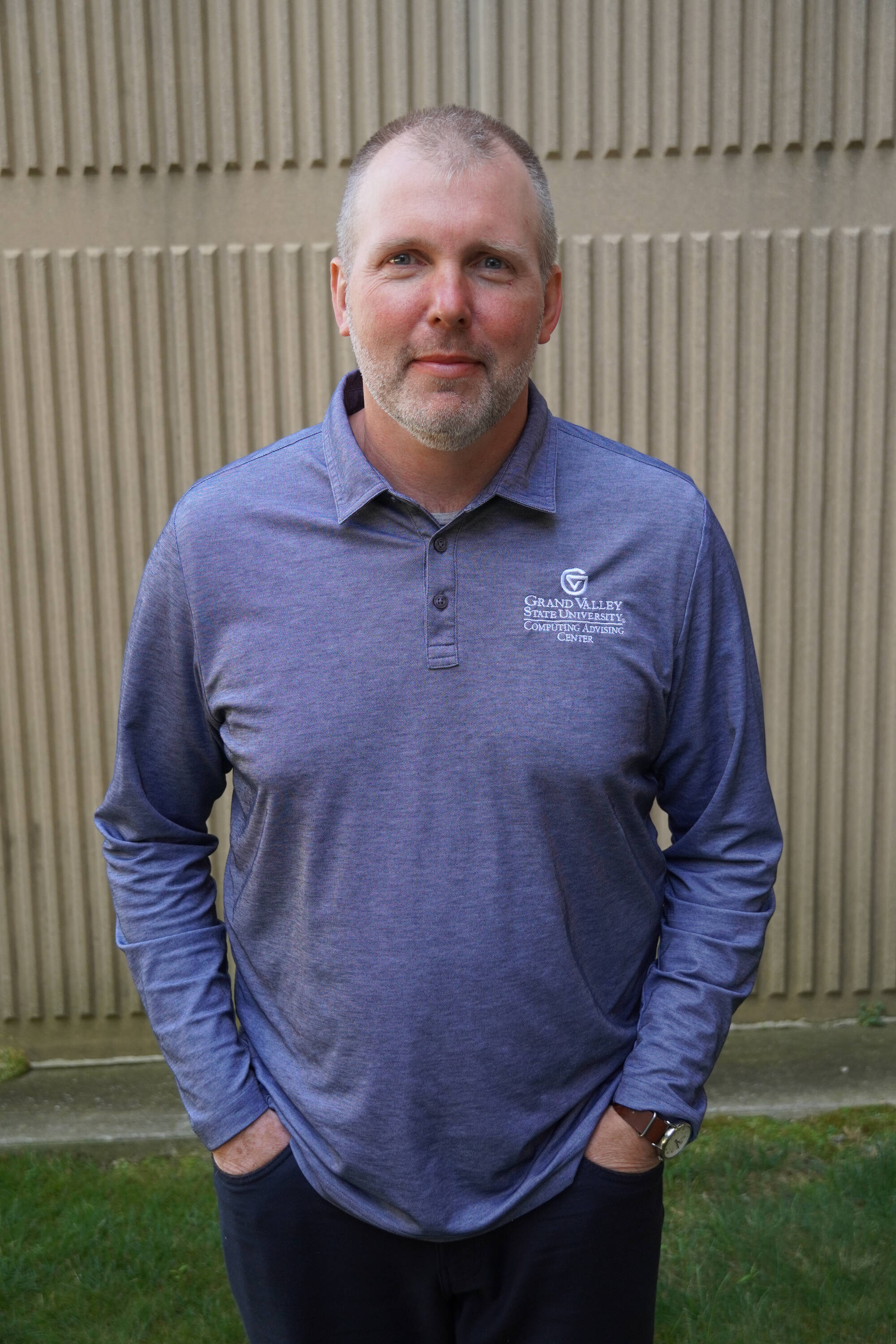Image of Colin DeKuiper smiling at the camera wearing a gray collared long sleeve standing in front of a textured tan background.